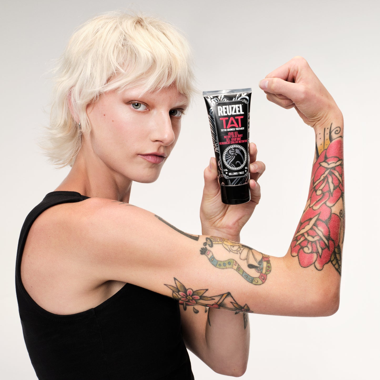 Person with tattoos holding a Reuzel VIVID Gel tattoo aftercare product in a tube.